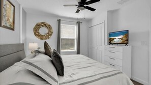 Ein Schlafzimmer mit einem Bett, einem Deckenventilator, einem Fernseher auf einer Kommode und einem Fenster mit Jalousien.