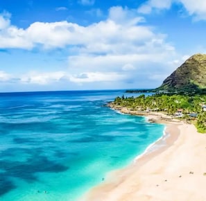 A coastal beach with clear turquoise waters, a sandy shore, and a small settlement with buildings and greenery. - 
Amazing Ocean Views - Beachfront 2BR with AC at Hawaiian Princess Makaha Resort (Waianae)