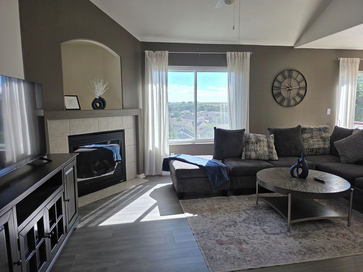 Ein modernes Wohnzimmer mit Kamin, einer Sofagarnitur, einem Fernseher und einem großen Fenster mit Vorhängen.