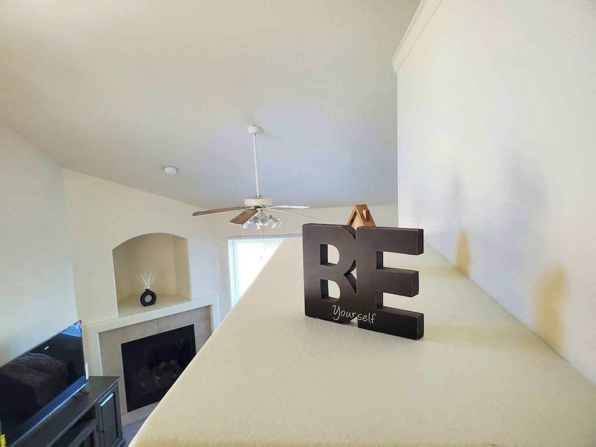 A modern living room with a fireplace, a ceiling fan, and a decorative sign that reads 'BE Yourself'.