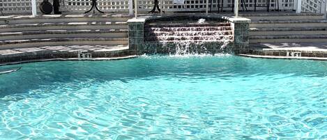 A clear pool with a waterfall feature, surrounded by a white fence with decorative fish and a pirate statue.