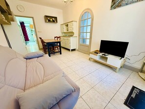 En stue med sofa, fjernsyn på et skærmstativ, spiseområde med stole og bord, samt et køkken med skabe.