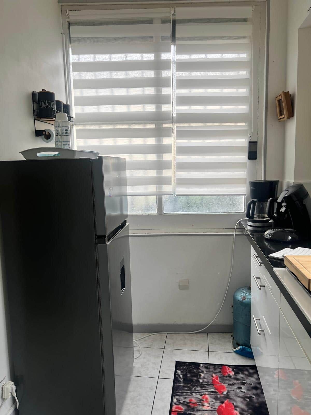 Een keuken met een koelkast, een koffiezetapparaat en een raam met jaloezieën.