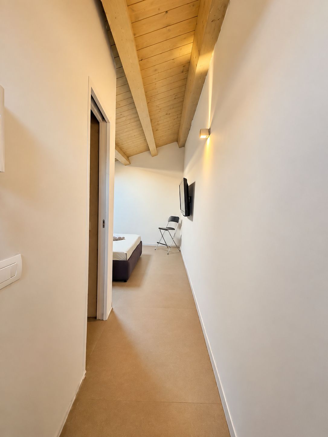 Un corridoio stretto con soffitto spiovente, un letto, una sedia e un televisore fissato al muro.