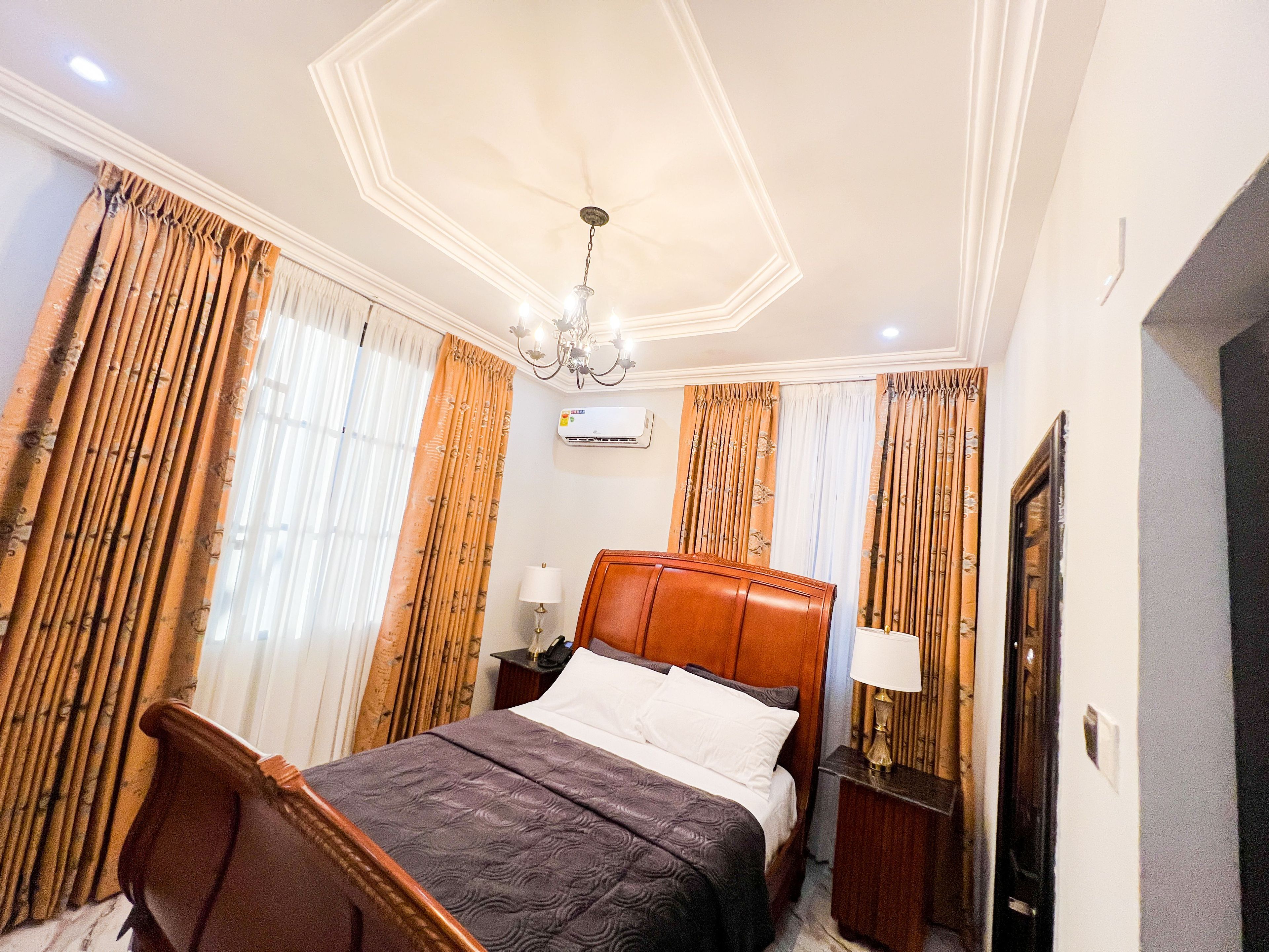 A hotel room with a bed, curtains, a chandelier, an air conditioning unit, and a nightstand.