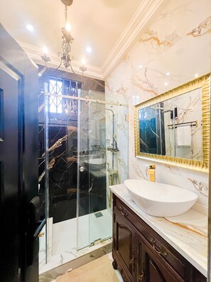 A bathroom with a glass-enclosed shower, a white sink, and a marble wall. - AfriBella Executive Suite (Kumasi)