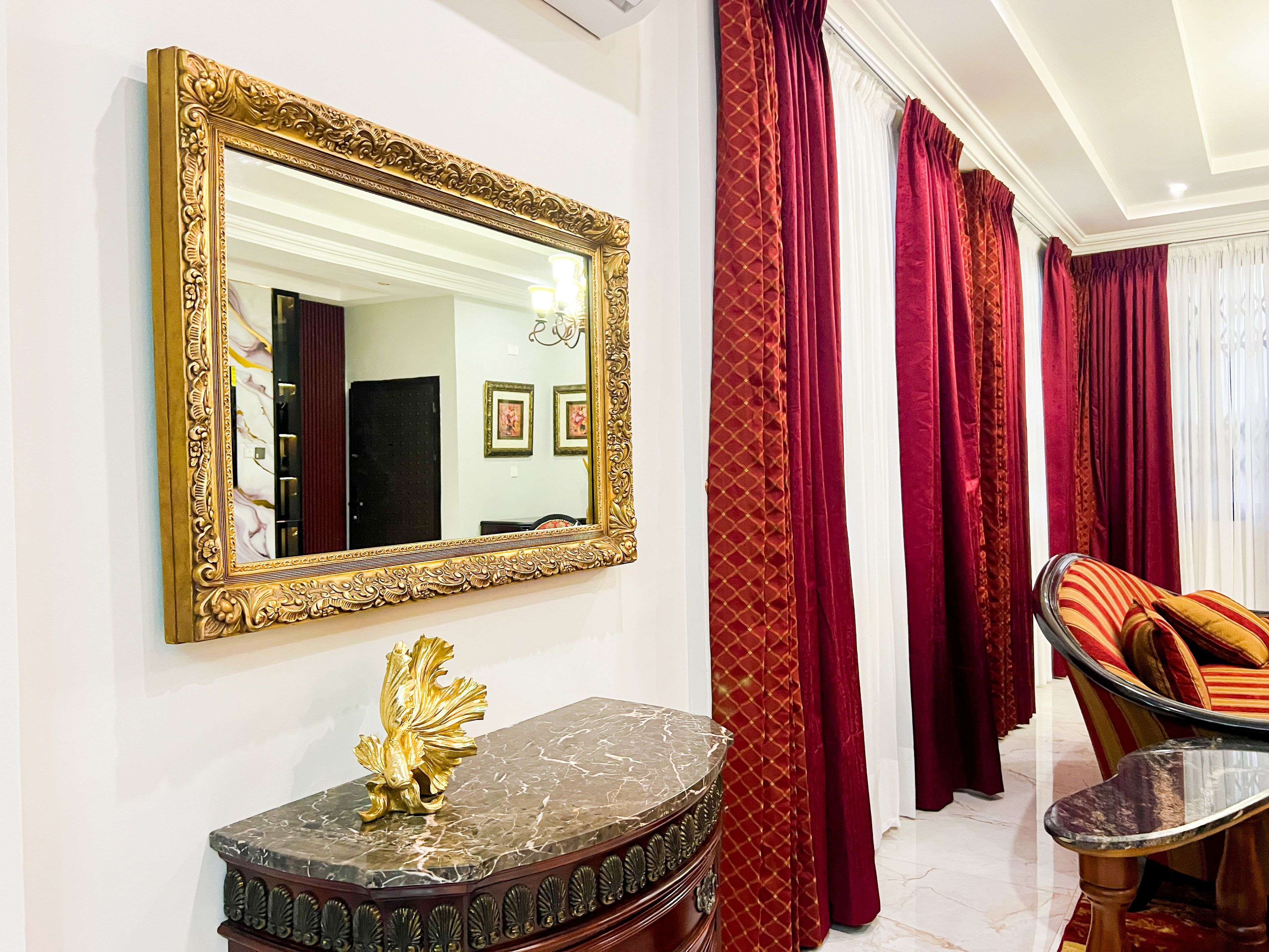 A room with a large ornate mirror, a marble console table with a golden lion sculpture, and red curtains.