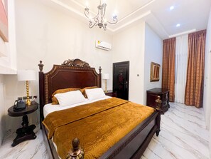 A hotel room with a large bed, a bedside table, a lamp, an air conditioning unit, a door, and a curtain. - AfriBella Executive Suite (Kumasi)