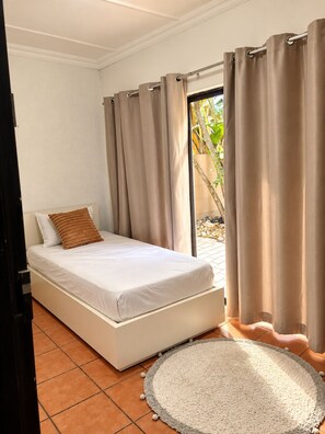 A single bed with a white mattress and a brown pillow, a round rug with a gray border, and a sliding glass door with beige curtains. - Perfect 3-bedroom house in Randburg for a relaxing stay (Randburg)