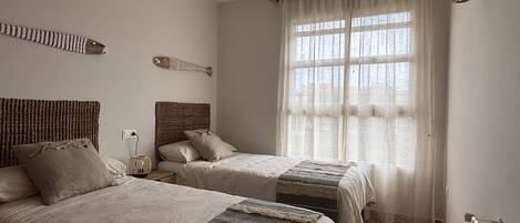 A bedroom with two beds, a large window with sheer curtains, and a radiator on the wall.