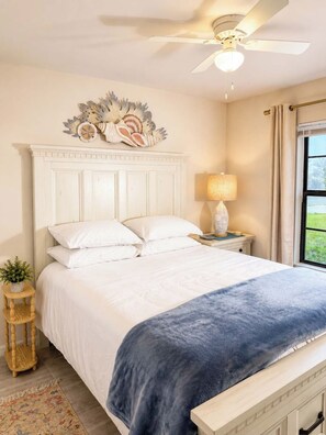 A bedroom with a bed, a ceiling fan, a nightstand with a lamp, a decorative wall piece, and a window with curtains. - 2025 Waterfront 3/2 Canal Views, Garage, Laundry (Cape Coral)