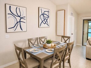 A dining area with a table set for four, surrounded by wooden chairs. There are two abstract paintings on the wall above the table. - 2025 Waterfront 3/2 Canal Views, Garage, Laundry (Cape Coral)