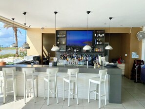 A bar area with a counter, white bar stools, and a large screen above the bar. - Luxury 3-Bedroom Retreat,Pools, Beaches & Snorkeling (Oranjestad)
