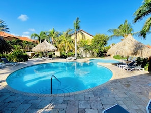 A swimming pool with lounge chairs and thatched umbrellas, surrounded by palm trees and a building with a red roof. - Luxury 3-Bedroom Retreat,Pools, Beaches & Snorkeling (Oranjestad)