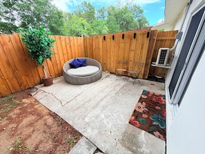 A backyard with a wooden fence, a round outdoor seating area with cushions, a small garden, and a door with a decorative mat. - Cozy Lodge Studio with Rustic Charm (Jacksonville)