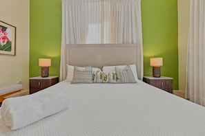 Una cama con edredón blanco, varias almohadas y una manta blanca. Hay dos mesitas de noche con lámparas. La habitación tiene paredes verdes y un ventanal con cortinas blancas.