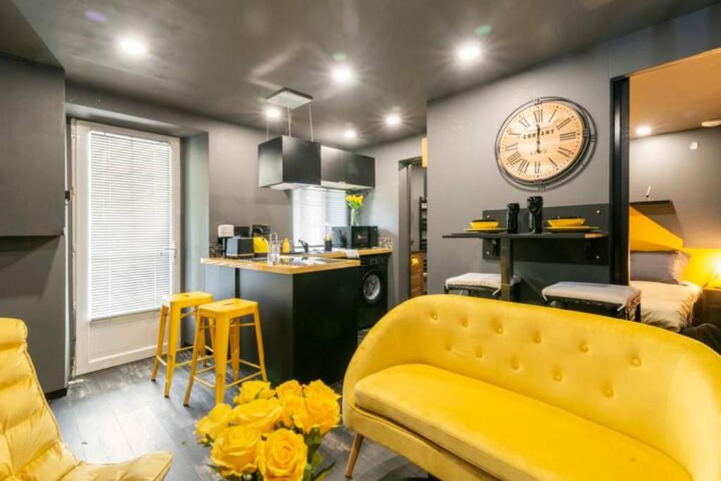 Un salon moderne avec un canapé jaune, un coin repas avec des tabourets et une cuisine ornée d’une horloge au mur.