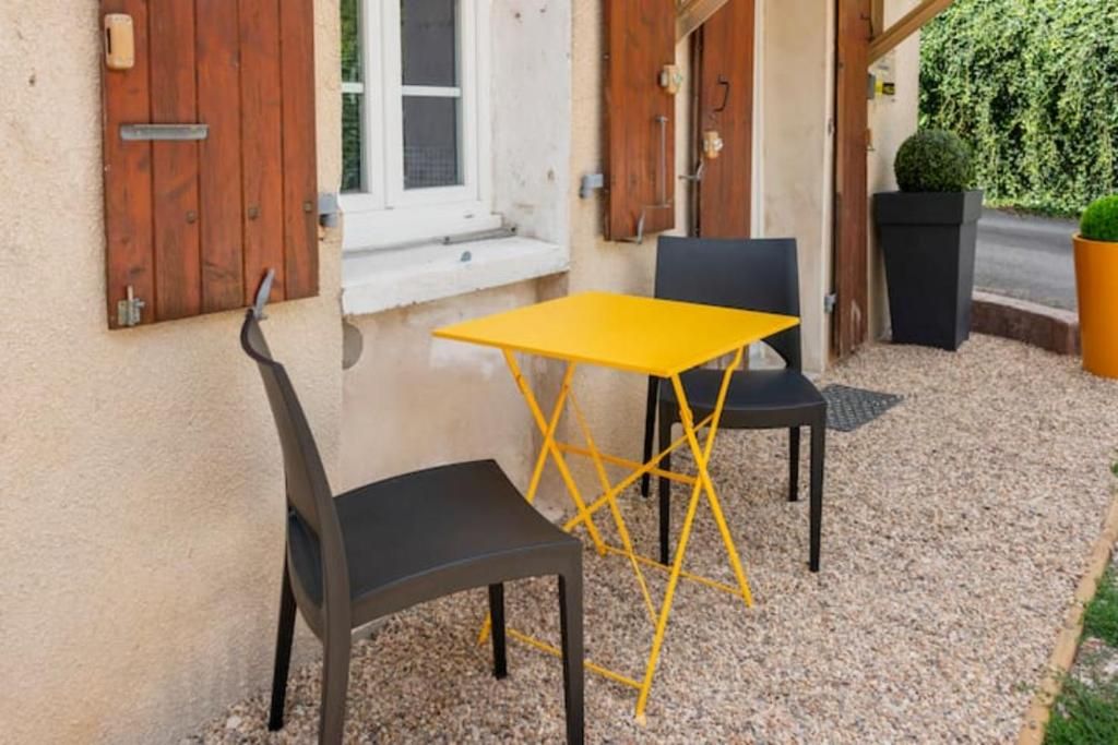 An outdoor seating area with a yellow table and two chairs, a wooden door, and a window with shutters.
