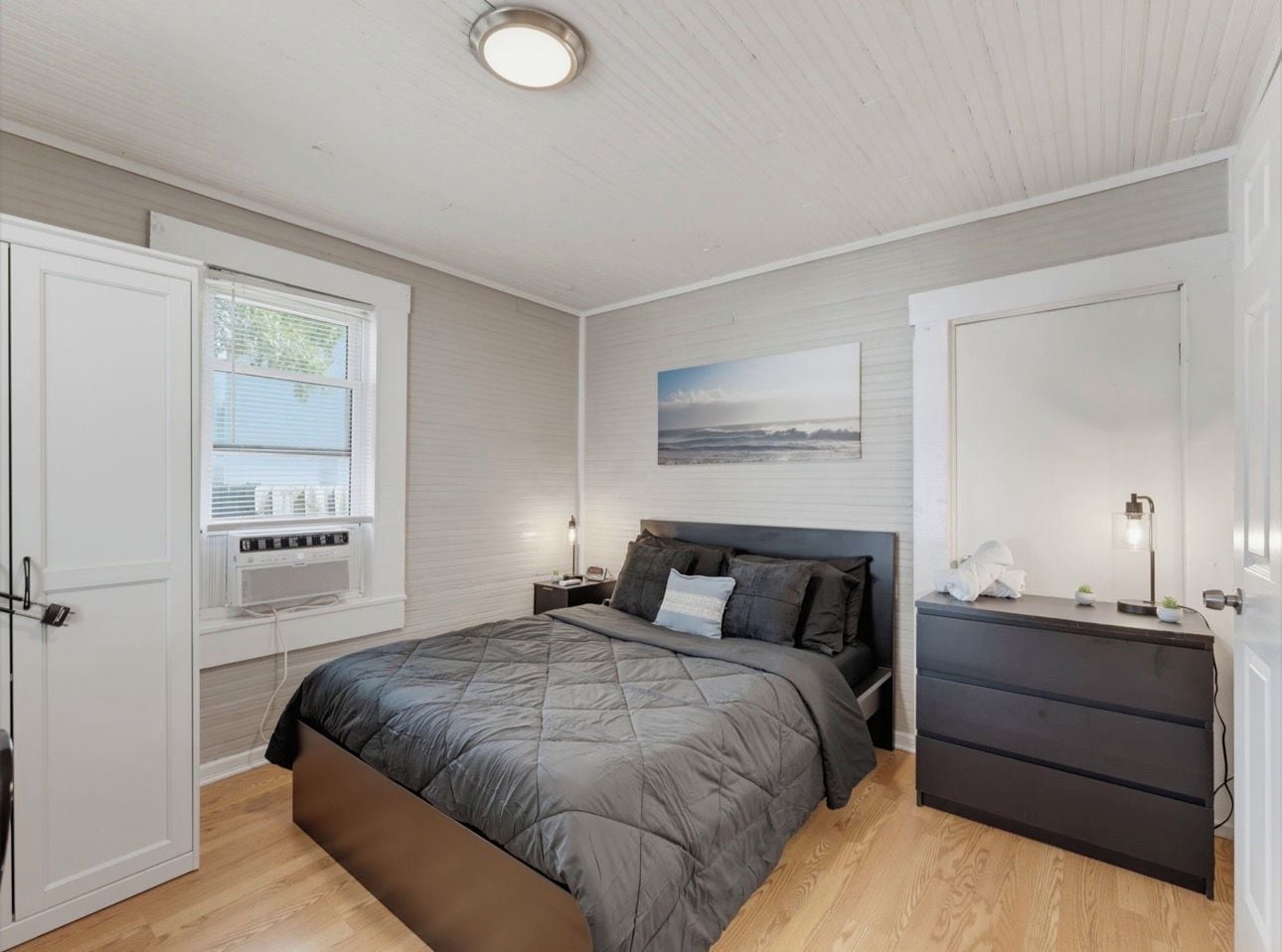 Un dormitorio con cama, mesitas de noche, cómoda, ventana y un cuadro en la pared.