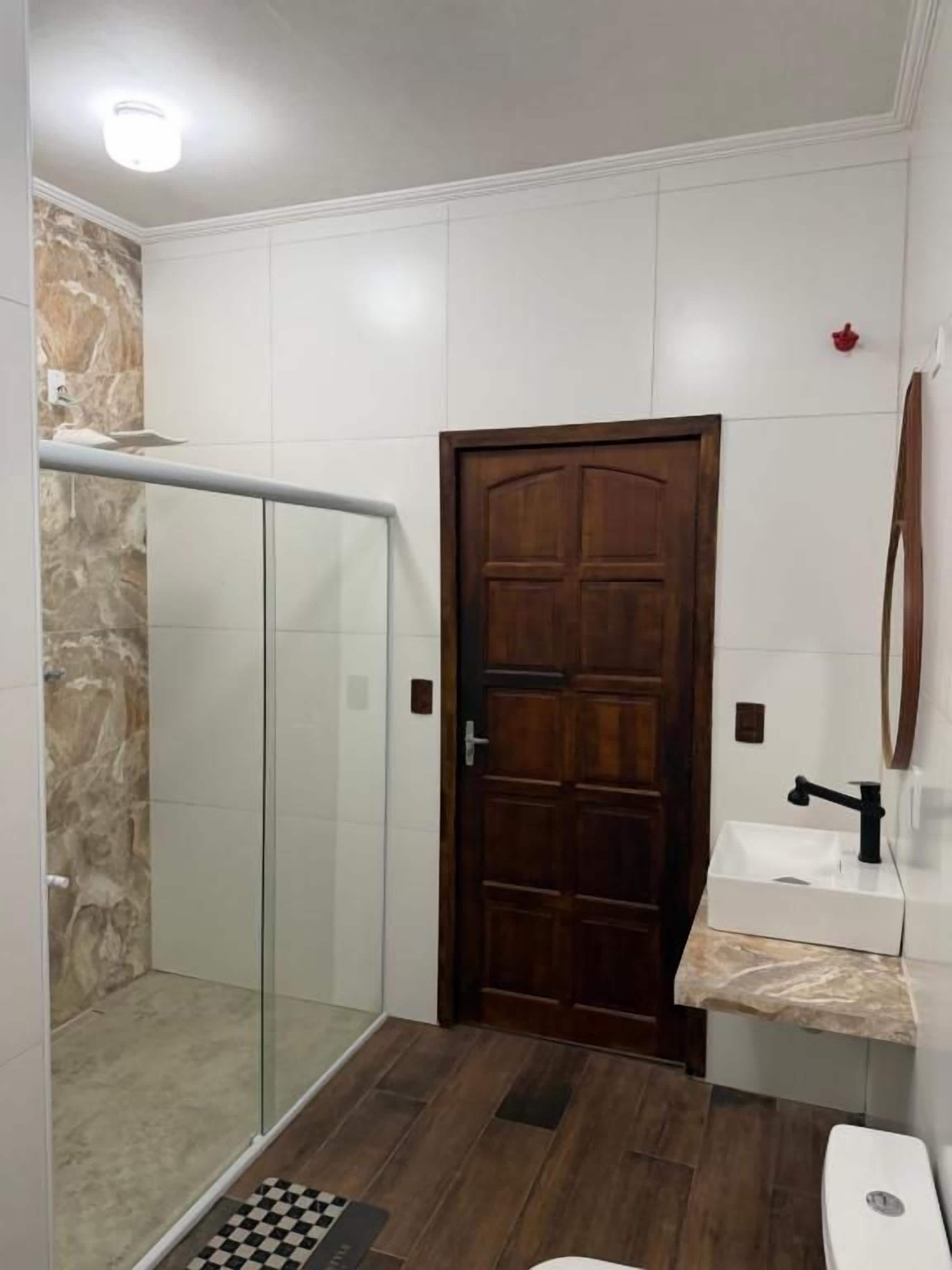 A bathroom with a glass shower enclosure, a wooden door, a sink with a marble countertop, and a toilet.