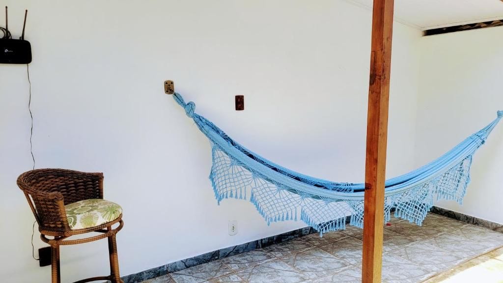 Un hamac tendu entre deux poteaux en bois, une chaise en osier avec un coussin, et un mur blanc avec une prise de courant.