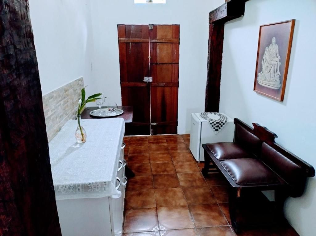 A small, compact room with a tiled floor, a white table with a vase, a bench, a wooden door, and a framed picture on the wall.