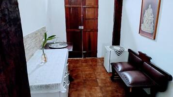 A small, compact room with a tiled floor, a white table with a vase, a bench, a wooden door, and a framed picture on the wall.