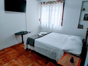 Un dormitorio con una cama, una mesita, un televisor montado en la pared y un ventanal con cortinas.