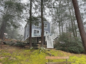 Eine zweistöckige Holzhütte mit einer Terrasse und Treppe, umgeben von einem Wald.