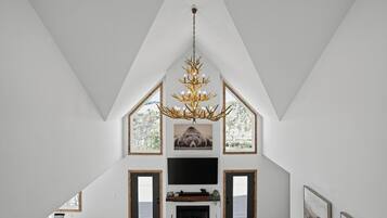 Amplia sala de estar con techo alto, chimenea, televisor y un ventanal con vistas a árboles.