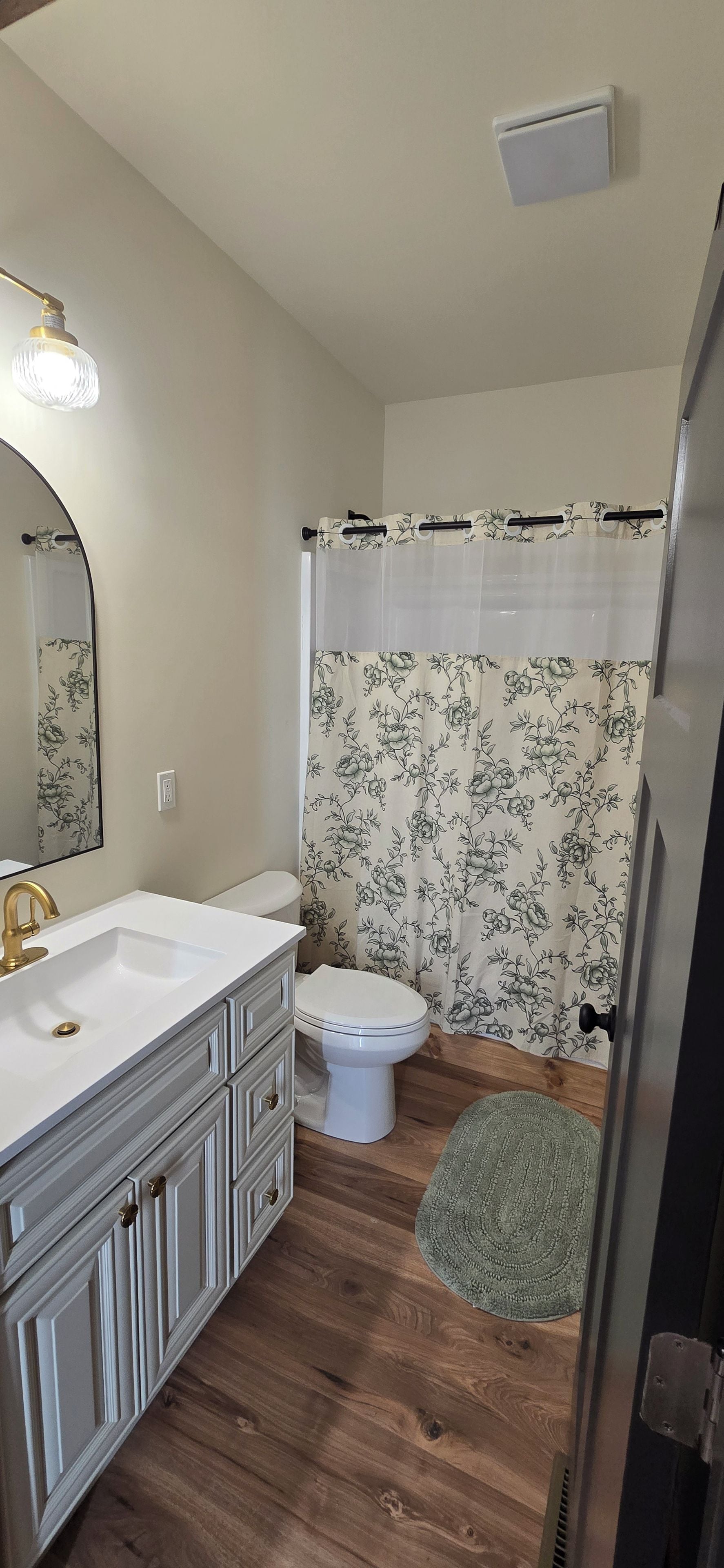 A bathroom with a white sink, a toilet, a shower curtain with a floral pattern, and a mirror with a light fixture.