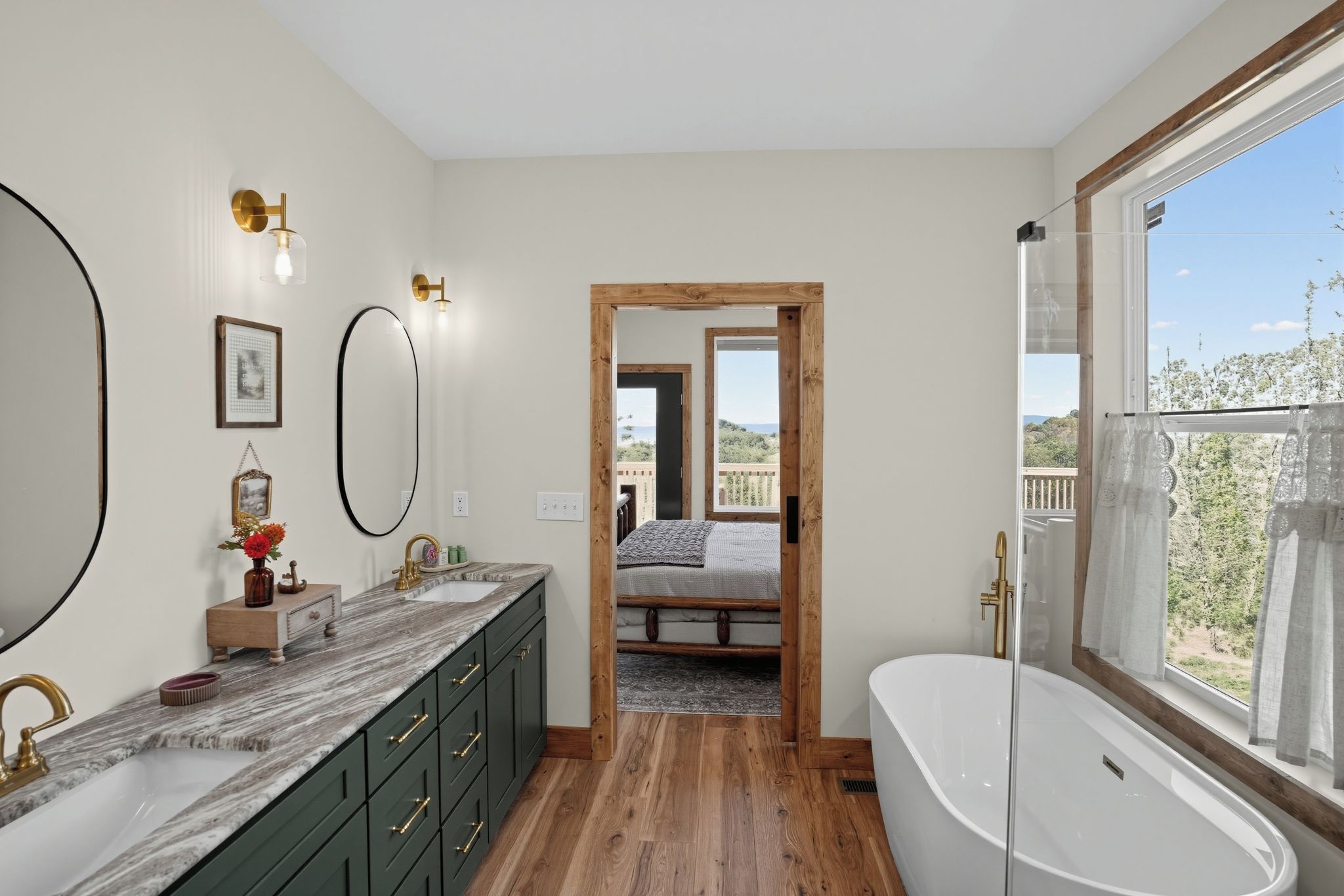 A modern bathroom with a large bathtub, a double sink vanity, and a large mirror. There is a wooden floor and a view of the outdoors through a window.