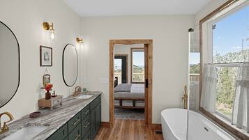 A modern bathroom with a large bathtub, a double sink vanity, and a large mirror. There is a wooden floor and a view of the outdoors through a window.