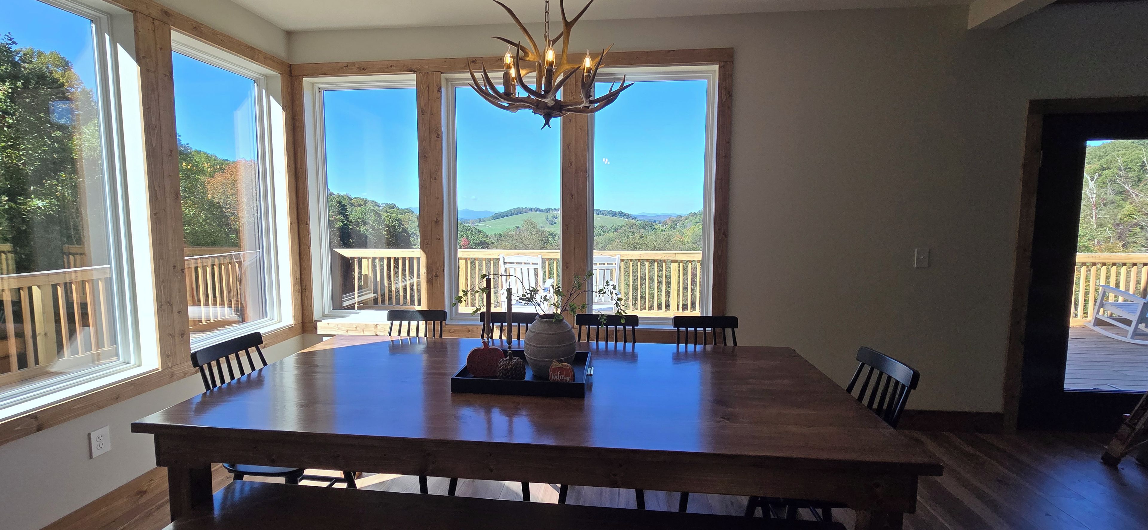 A dining area with a large wooden table, six chairs, a chandelier, and a view of a deck and landscape through large windows.