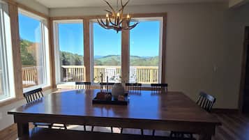 A dining area with a large wooden table, six chairs, a chandelier, and a view of a deck and landscape through large windows.