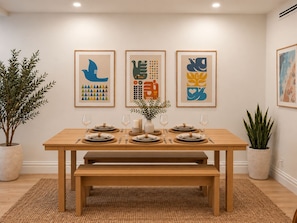 Un comedor con una mesa de madera dispuesta para cuatro personas, una obra de arte enmarcada en la pared y plantas en macetas.