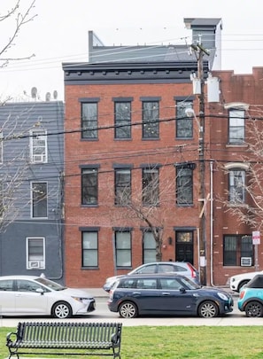 A multi-story building with a brick facade, several windows, and a parking lot with cars. - NYC Skyline Rooftop · 3BR Row House · Sleeps 9 (Jersey city)