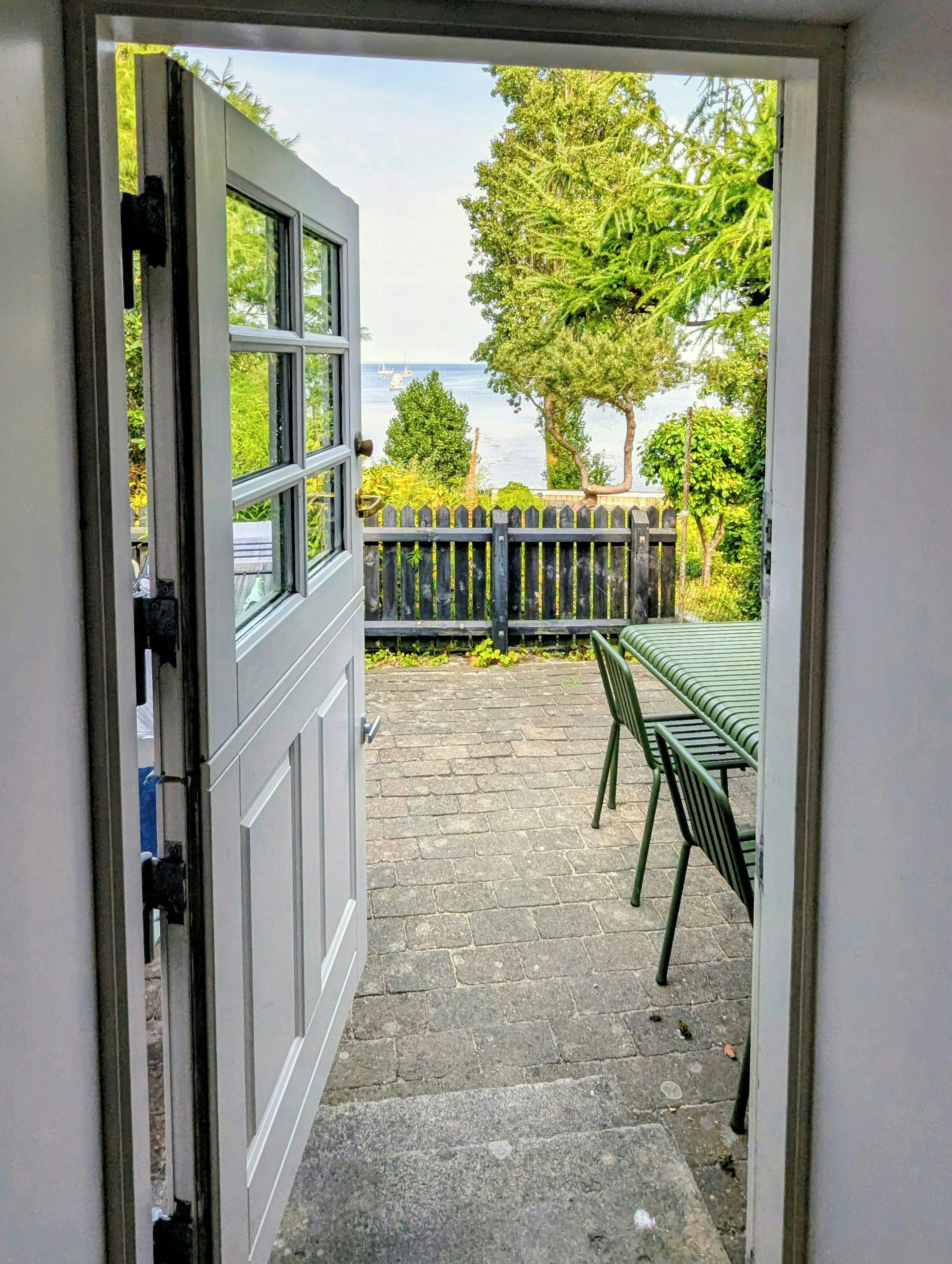 An open door leading to a patio with a table and chairs, a wooden fence, and a view of the sea.
