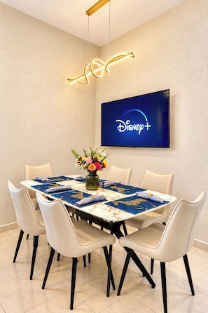 A dining area with a table set for six, a flat-screen TV on the wall displaying the Disney+ logo, and a modern light fixture above the table. - Villa Morán - El Almendro offers luxury, safety, and serenity. All new for you! (Sonsonate)