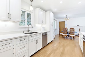 A modern kitchen with white cabinetry, a built-in dishwasher, and a dining area with a wooden table and chairs.