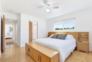 A bedroom with a bed, bedside tables, a ceiling fan, a window, and a door leading to another room.