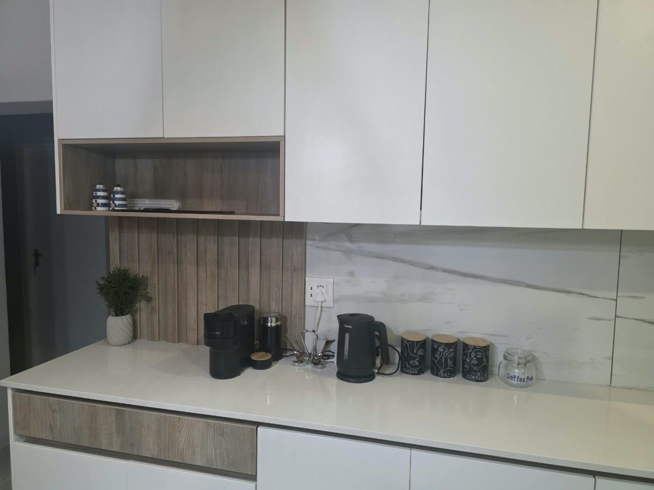 A modern kitchen with white cabinets, a marble backsplash, and a countertop with coffee-making equipment and mugs.
