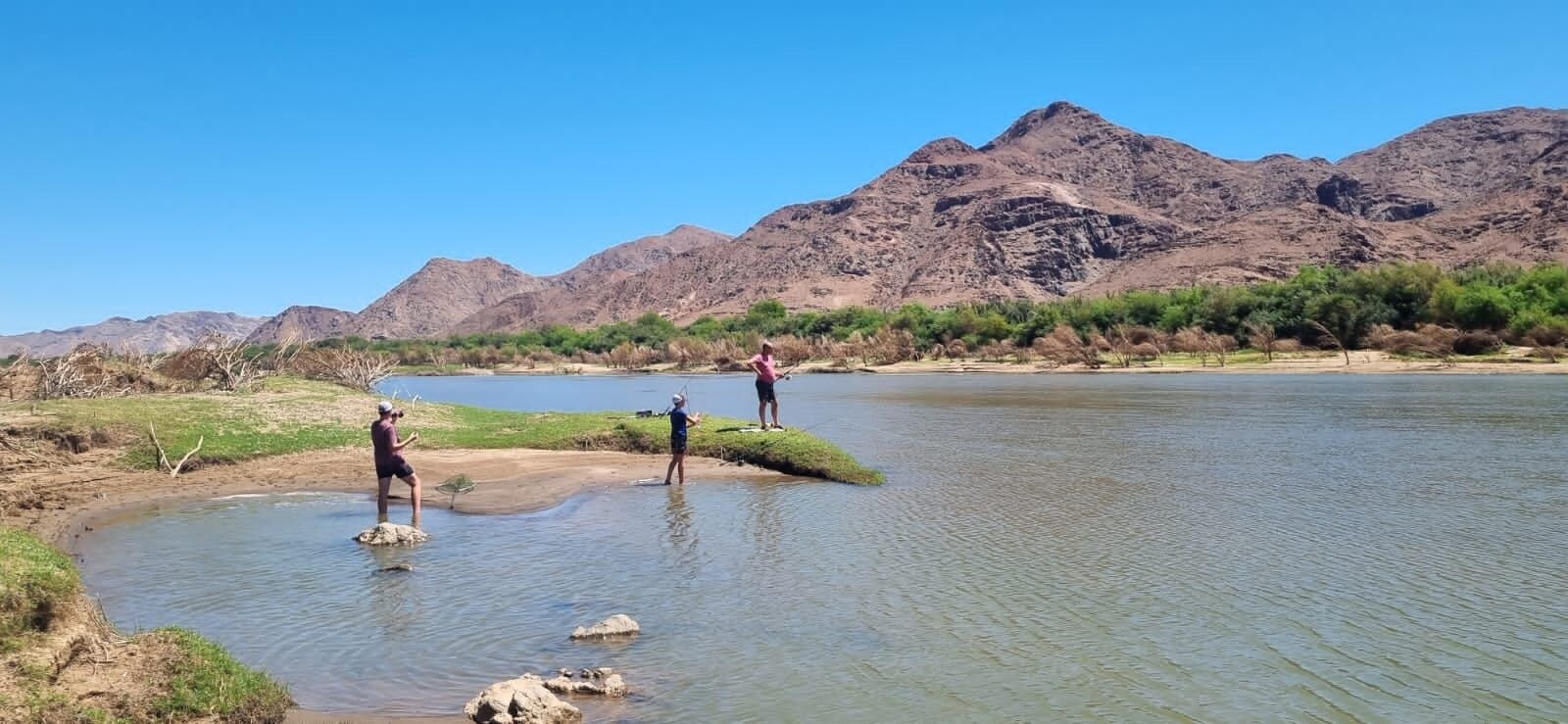 Une rivière où des personnes se tiennent dans l’eau, entourées de montagnes et d’une végétation clairsemée.