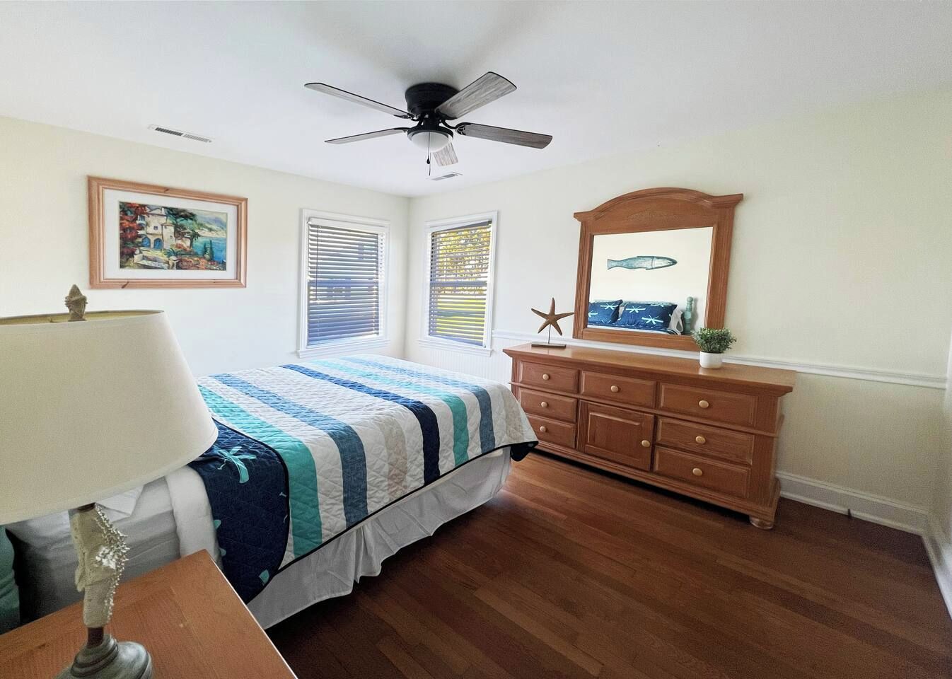 A bedroom with a bed, dresser, ceiling fan, and artwork on the walls.