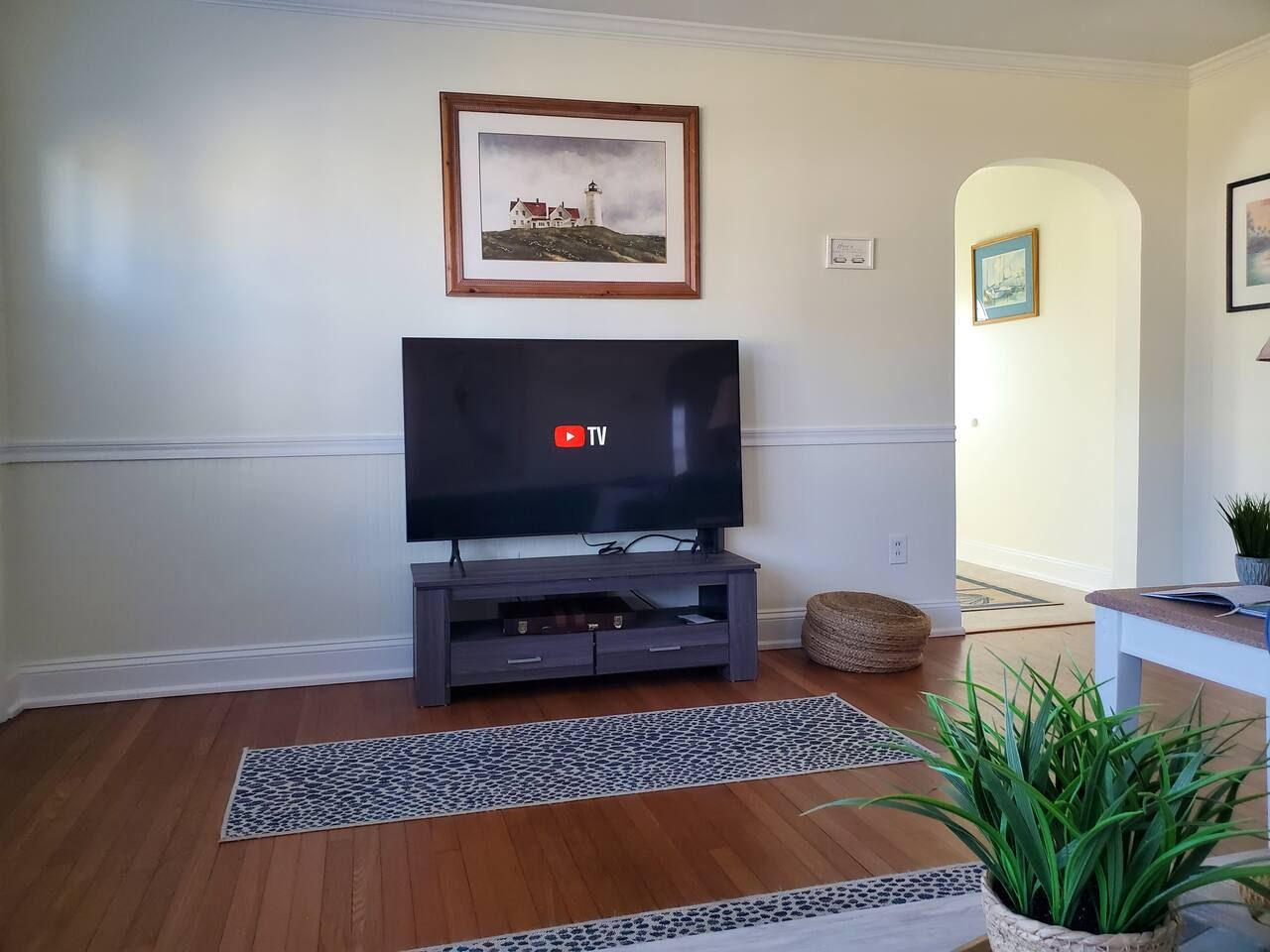 A living room with a flat-screen TV on a wooden stand, a framed painting above it, and a potted plant on the floor.