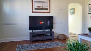A living room with a flat-screen TV on a wooden stand, a framed painting above it, and a potted plant on the floor.