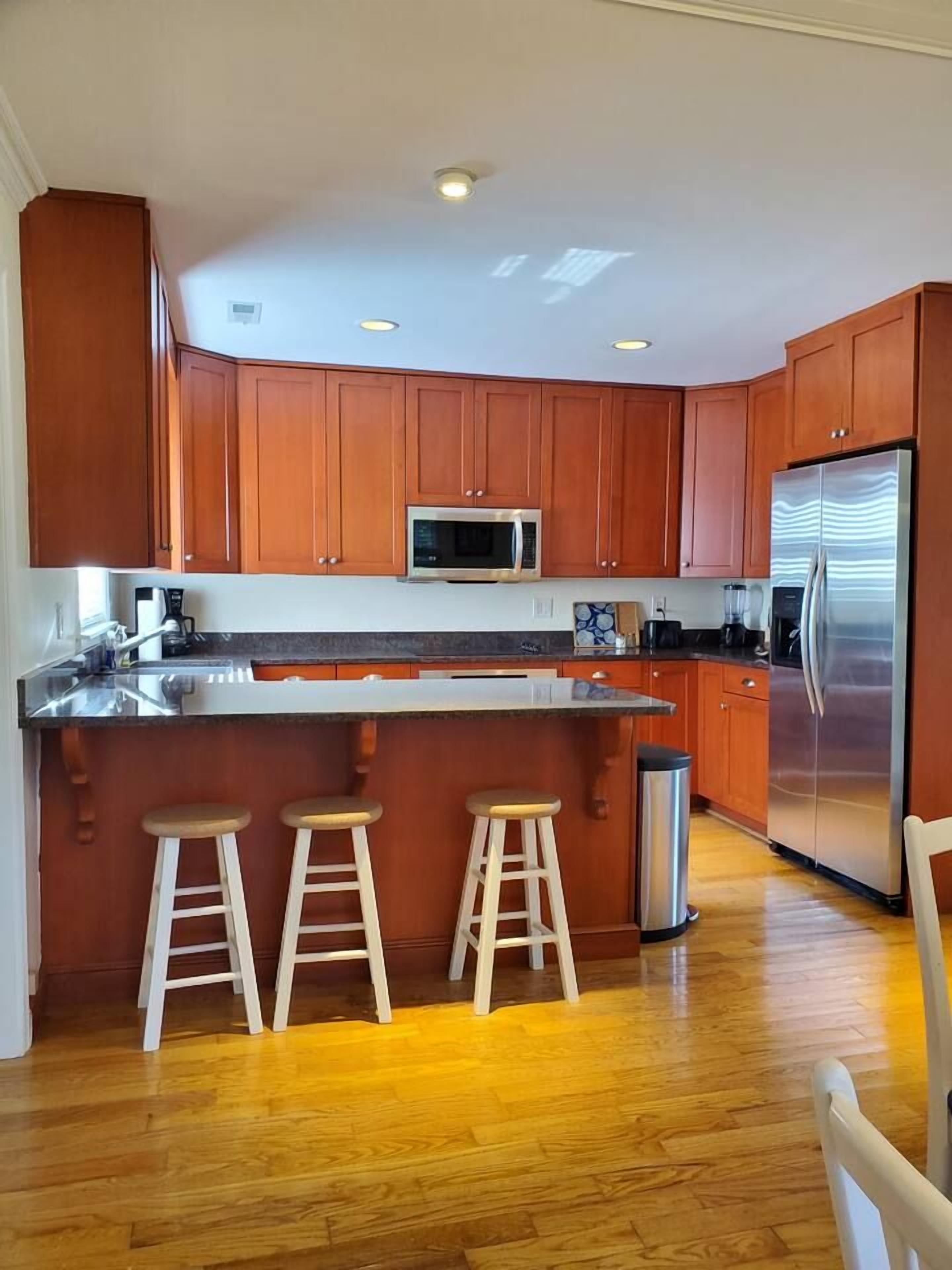A kitchen with wooden cabinets, a stainless steel refrigerator, a microwave, and a bar stool area.