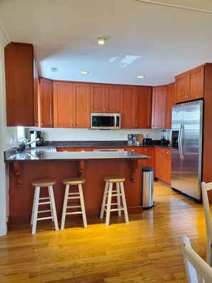 A kitchen with wooden cabinets, a stainless steel refrigerator, a microwave, and a bar stool area.