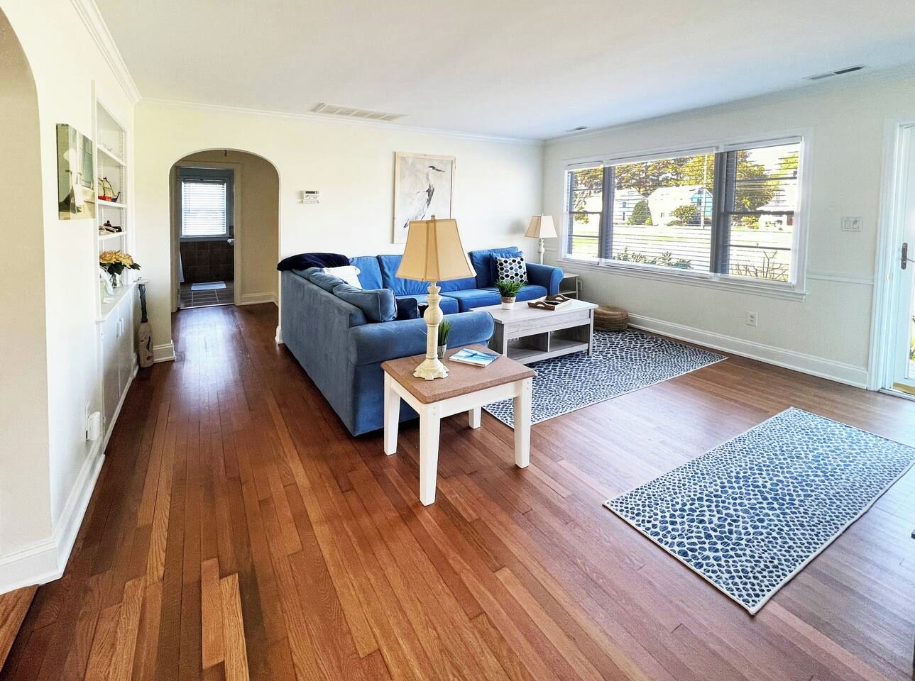 Een woonkamer met houten vloeren, een blauwe bank, een klein tafeltje met een lamp en een groot raam met uitzicht.