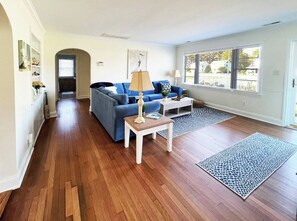 A living room with hardwood flooring, a blue sofa, a small table with a lamp, and a large window with a view.